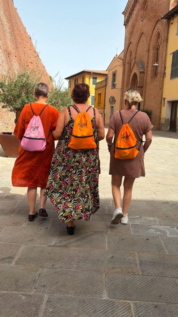Tre kvinnor med färgglada gruppreseryggsäckar strosar i en gammal italiensk by, kvinnor, solsken.