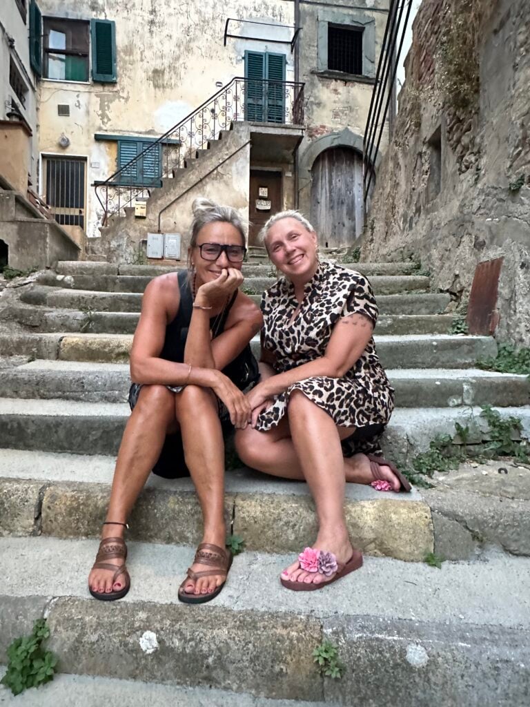 Två blonda kvinnor i sandaler sitter på en stentrappa i en gammal stad och ler.