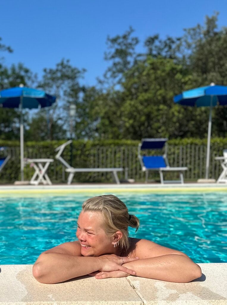 En blond kvinna hänger med armarna på poolkanten och kroppen i vattnet.