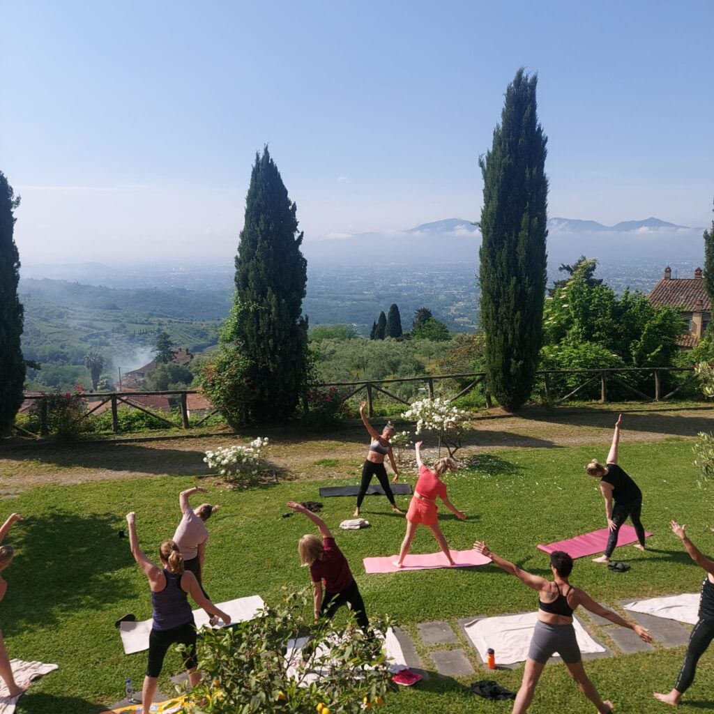 Ett gäng kvinnor gör yoga på en solig gräsmatta med utsikt över svindlande landskap.
