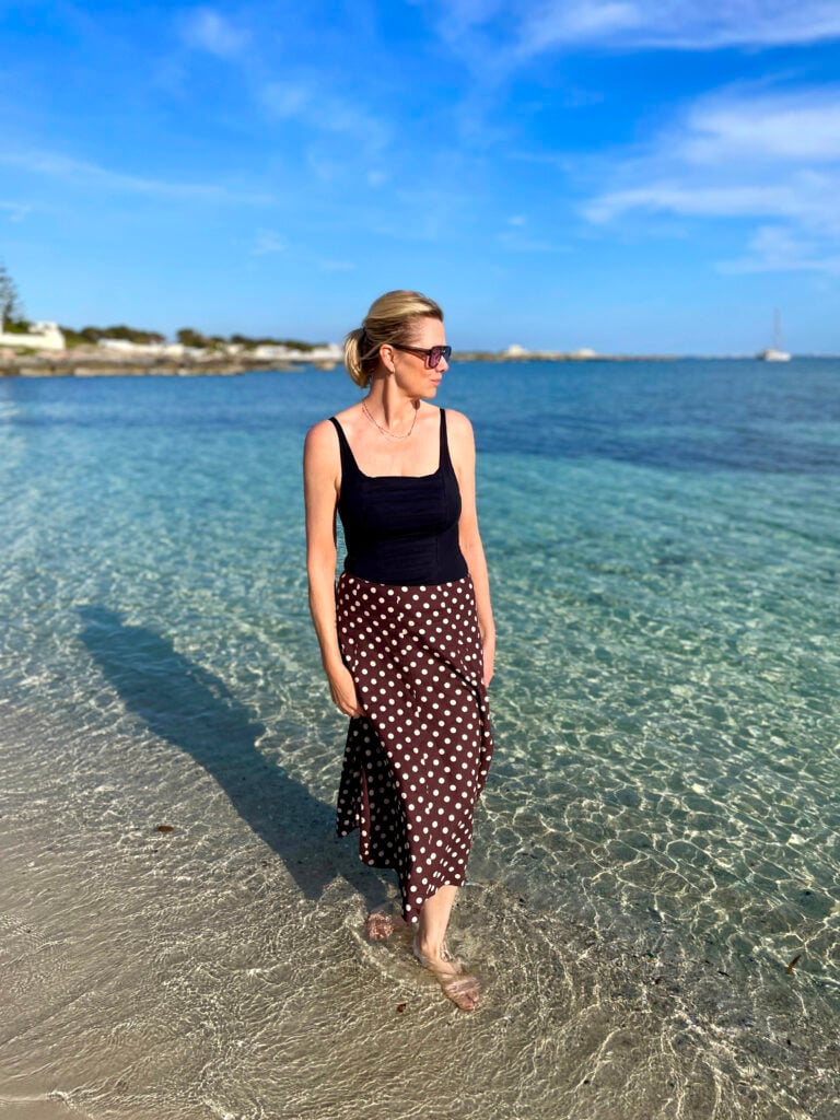 En kvinna i prickig kjol står med fötterna i vattenbrynet på en strand.