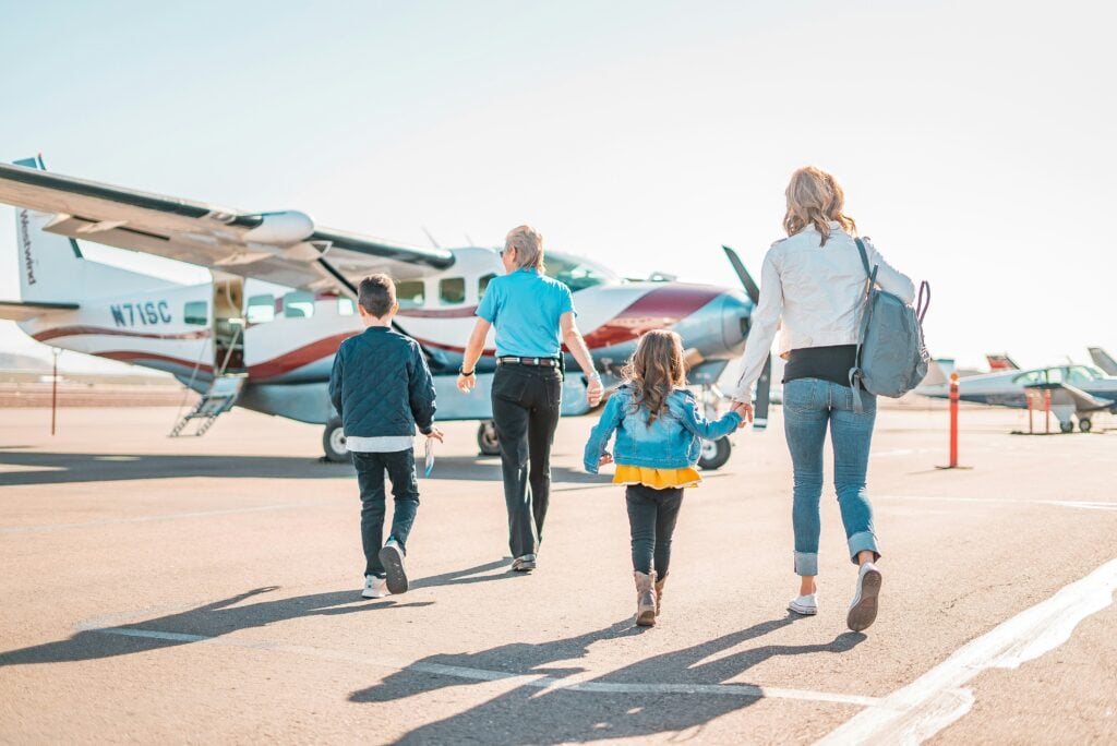 En familj på två vuxna och två barn går i solen över flygplatsen till ett flygplan.