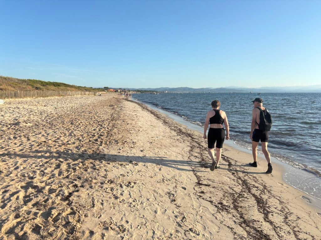 Två personer promenerar i shorts på en strand i vattenbryn. 