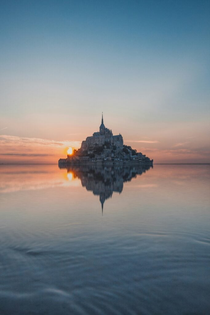 En toppig bebyggd ö syns i ett spegelblankt hav som reflekterar en solnedgång i pastellfärger. 