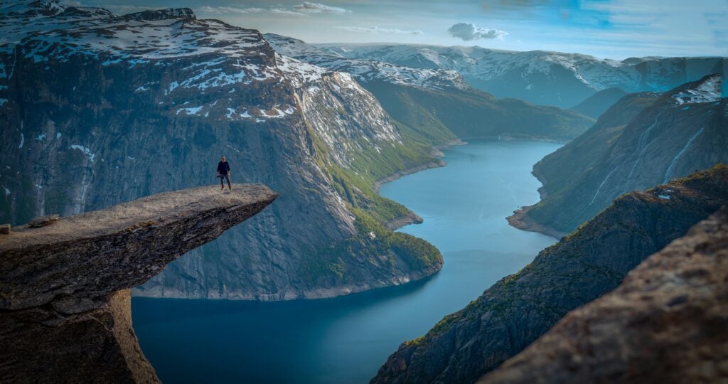 En man står på en klippa som sticker ut över en fjord bredvid dramatiska fjällsidor. 