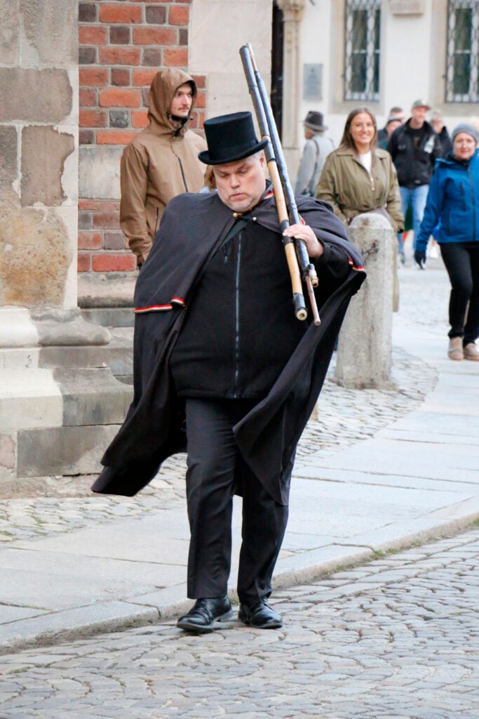 En svartklädd man i cape och svart stormhatt promenerar längs med kullerstensgatan med två långa stavar på axeln.