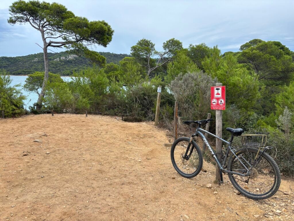 En cykel står parkerad mot en stolpe på en sandig brun grusväg, gröna träd och havet syns i bakgrunden. 