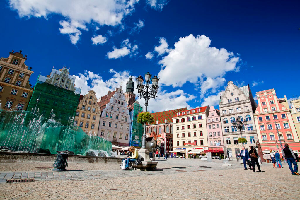 Solen skiner på ett klassiskt torg med en fontän och färgglada gamla stadshus. Människor flanerar.