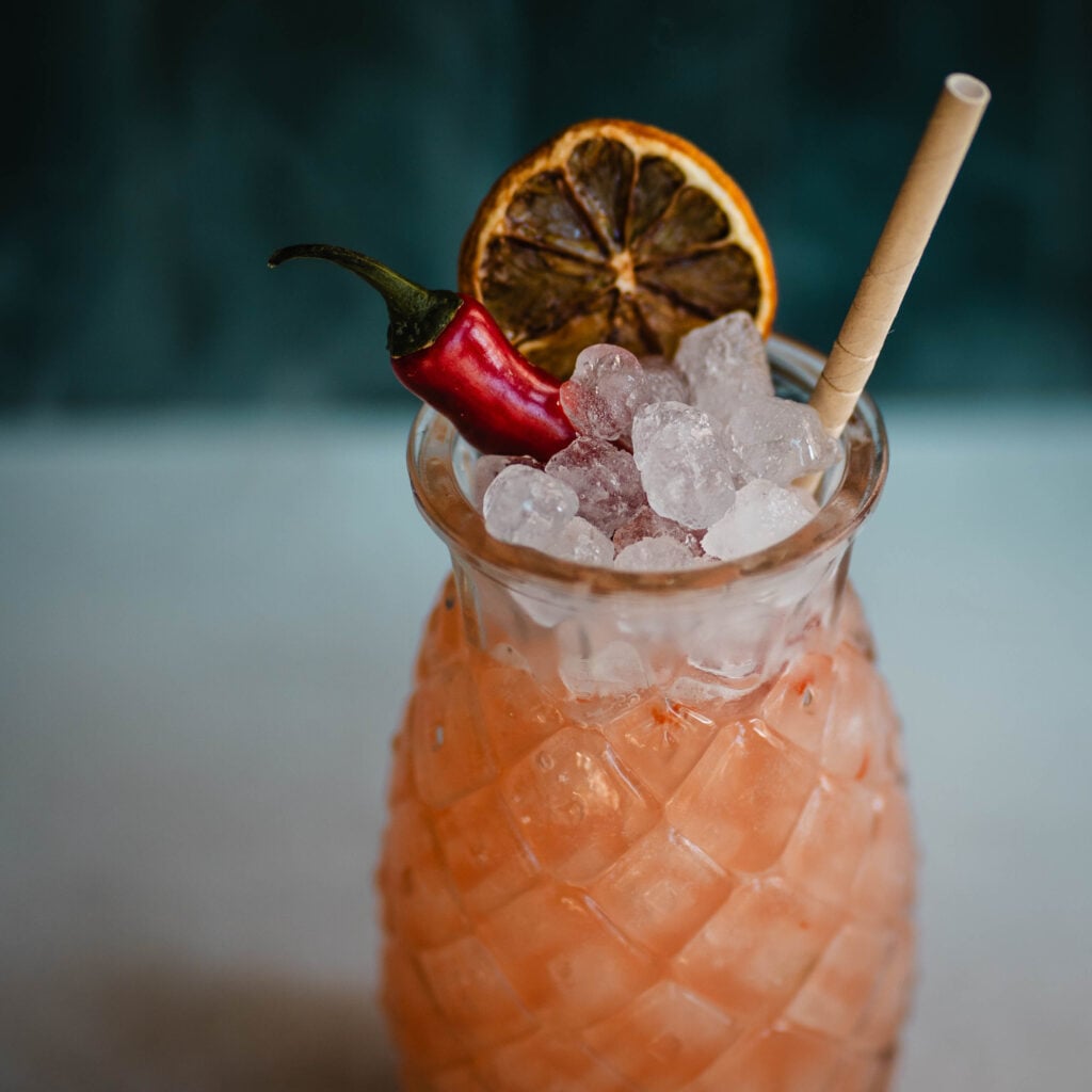 En orange drink i ett glas med is, chili, apelsinskiva, sugrör.