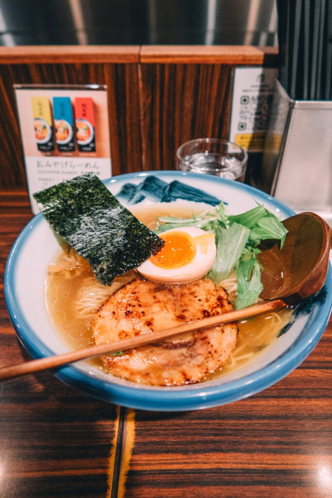 En rund skål med ramen, ett kokt ägg i, en djup slevliknande träslev.